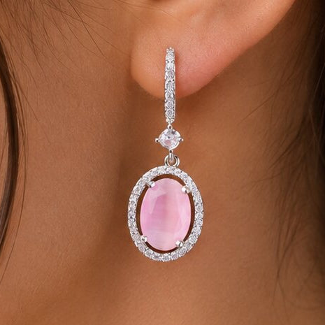 A close-up of a model's ear wearing a dangling earring with a pink oval gemstone surrounded by a silver-toned halo and small clear stones above it.