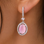 A close-up of a model's ear wearing a dangling earring with a pink oval gemstone surrounded by a silver-toned halo and small clear stones above it.