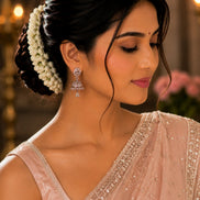 A woman in profile wearing a light pink embellished sari and matching blouse, with a white floral hair accessory and dangling earrings, against a softly blurred indoor background.