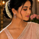 A woman in profile wearing a light pink embellished sari and matching blouse, with a white floral hair accessory and dangling earrings, against a softly blurred indoor background.
