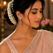 A woman in profile wearing a light pink embellished sari and matching blouse, with a white floral hair accessory and dangling earrings, against a softly blurred indoor background.