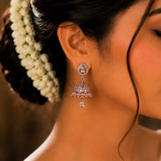 A close-up of a woman's ear wearing a dangling silver-toned jhumka earring adorned with clear stones, with dark hair and a white floral hair accessory visible against a warm brown background.