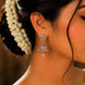 A close-up of a woman's ear wearing a dangling silver-toned jhumka earring adorned with clear stones, with dark hair and a white floral hair accessory visible against a warm brown background.