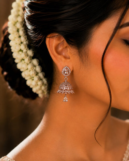 A close-up of a woman's ear wearing a dangling silver-toned jhumka earring adorned with clear stones, with dark hair and a white floral hair accessory visible against a warm brown background.